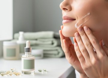 Close-up of a person applying a clear lotion to the face with skincare products nearby on a clean bathroom counter.