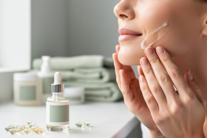 Close-up of a person applying a clear lotion to the face with skincare products nearby on a clean bathroom counter.